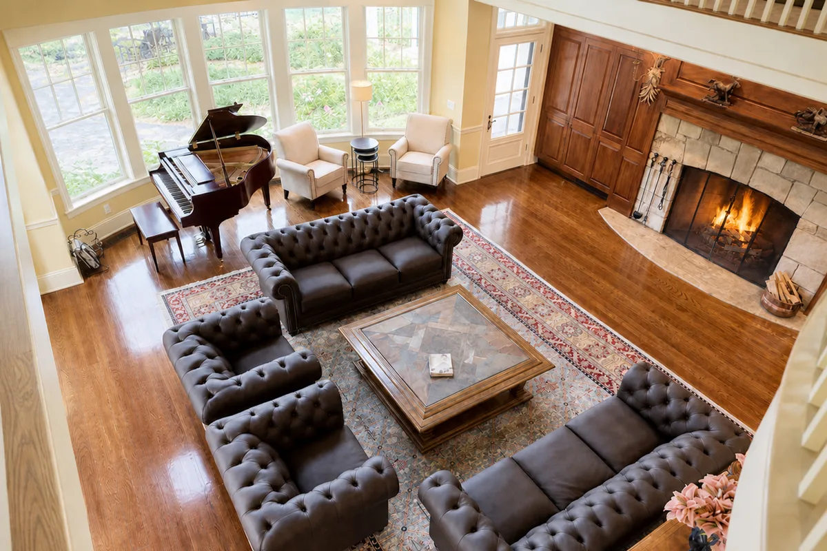 High-angle interior view looking down from the loft walkway onto the Great Room's spacious conversation seating area. Features two button-tufted grey Chesterfield sofas and two matching button-tufted armchairs arranged on a large red-patterned Persian rug around a square coffee table. A mahogany grand piano sits near large windows looking out at green gardens. Highlights polished hardwood floors and the edge of a massive stone fireplace. [Season: Summer] [Lighting: Bright, natural daytime light] [Content: Great room interior, furniture, piano, Persian rug, hardwood floors]