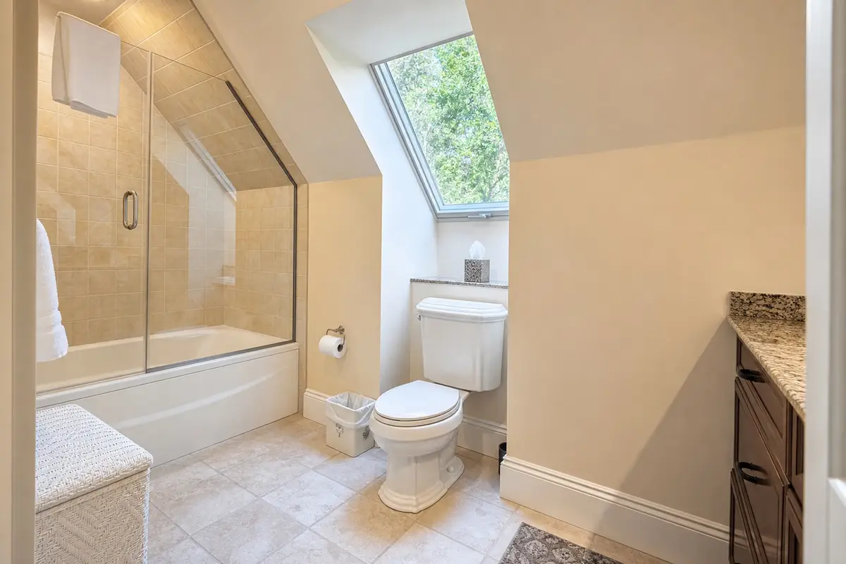 Ensuite guest bathroom under a sloped ceiling, showing a white toilet, a walk-in bathtub/shower with a glass door, a skylight letting in daylight, and dark wood vanity cabinets. [Season: Summer/Spring] [Lighting: Soft daylight and warm interior light] [Content: Guest bathroom, skylight, toilet, bathtub, vanity]