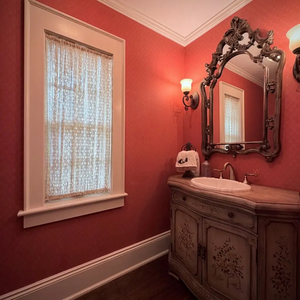 Half bath or powder room featuring deep reddish-pink walls, a white window with a lace curtain, an ornate dark-framed mirror, a classic wooden vanity with an undermount sink, and a wall sconce. [Season: Indoor] [Lighting: Warm, dim ambient light] [Content: Powder room, red walls, lace curtain, antique vanity, mirror]