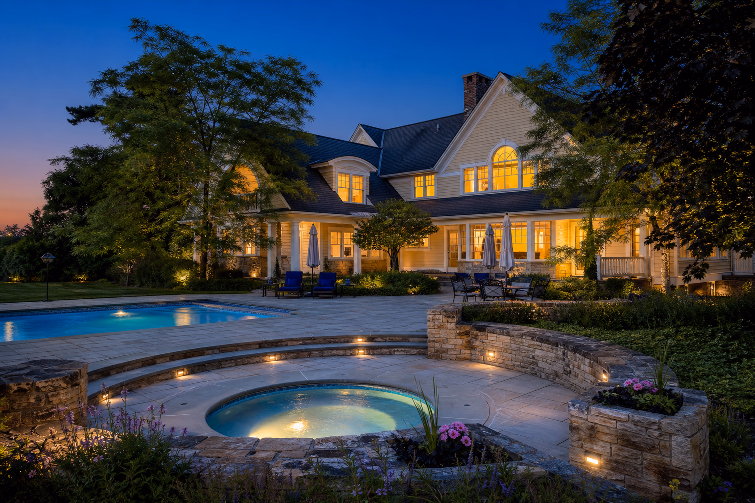 Rear exterior twilight view of the estate, showcasing the glowing warm windows overlooking the stone-paved patio and private swimming pool.