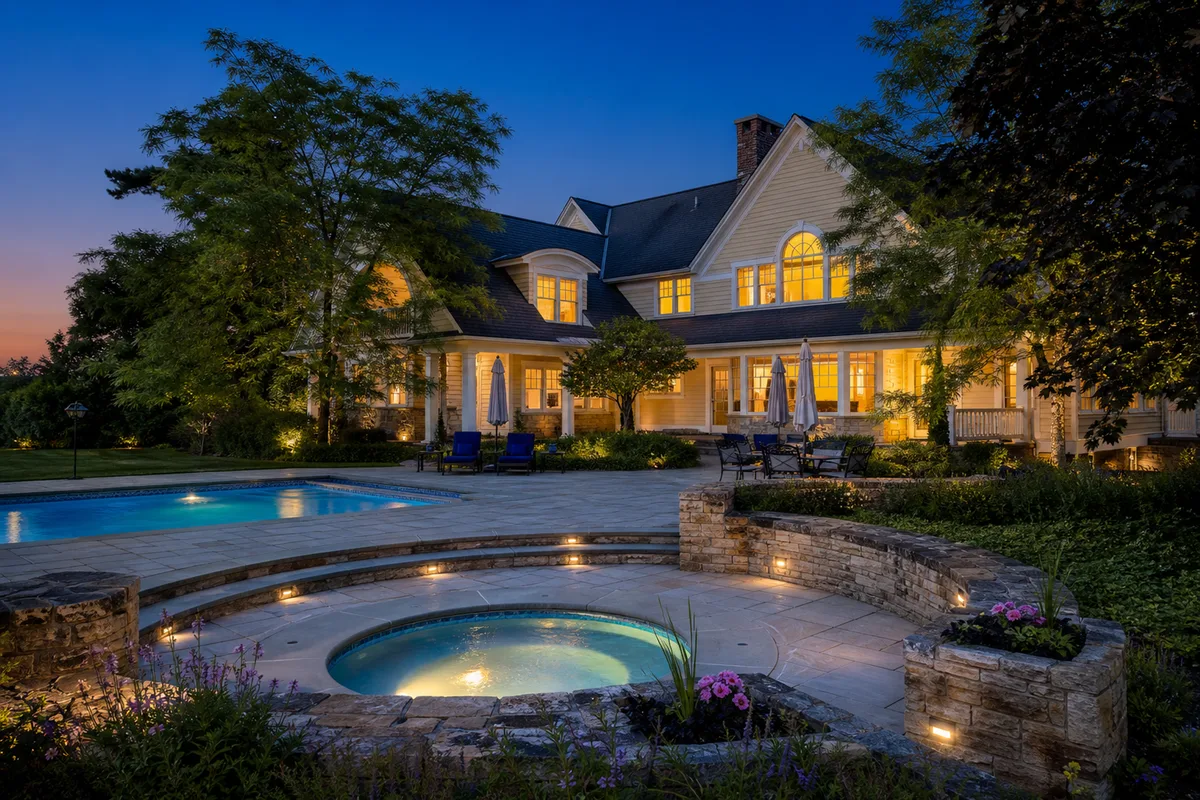 Dusk/twilight view of the rear of the estate house, highlighting the glowing warm windows. In the foreground is a stone-paved patio with blue lounge chairs next to a swimming pool, surrounded by green lawns and trees. [Season: Summer] [Lighting: Warm twilight / dusk] [Content: Backyard elevation, swimming pool, stone patio, twilight exterior]