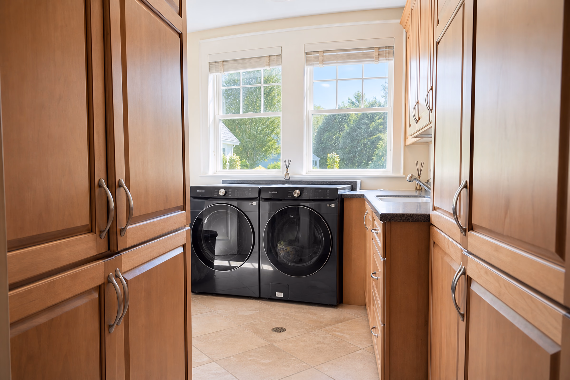 Spacious, high-end laundry room showing premium custom wood cabinetry on both sides, a tiled floor, and two modern dark grey front-load smart washers/dryers built into the cabinets under a window. [Season: Summer] [Lighting: Bright daylight from the window] [Content: Laundry room, washer and dryer, custom wood cabinets, folding counter]